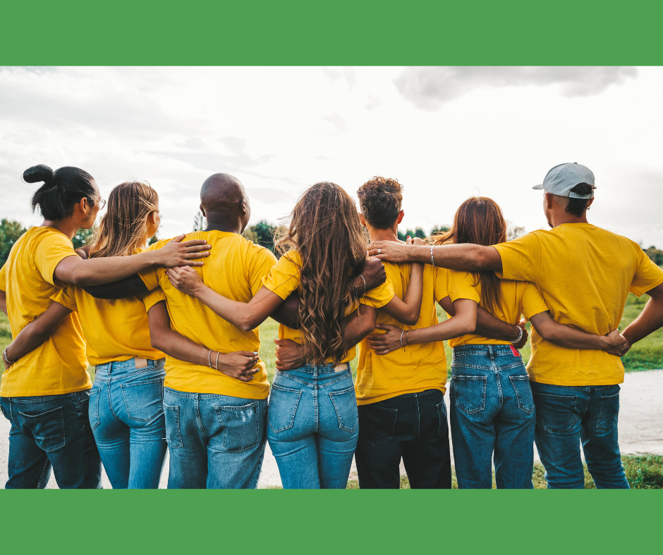 Group of seven diverse young adults in yellow shirts standing side by side with arms around each other outdoors near a body of water.