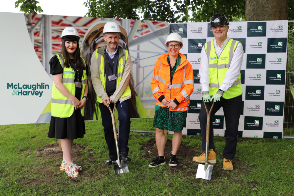 Four professionals in safety gear hold shovels at a ground breaking event on grass with promotional branding behind them.