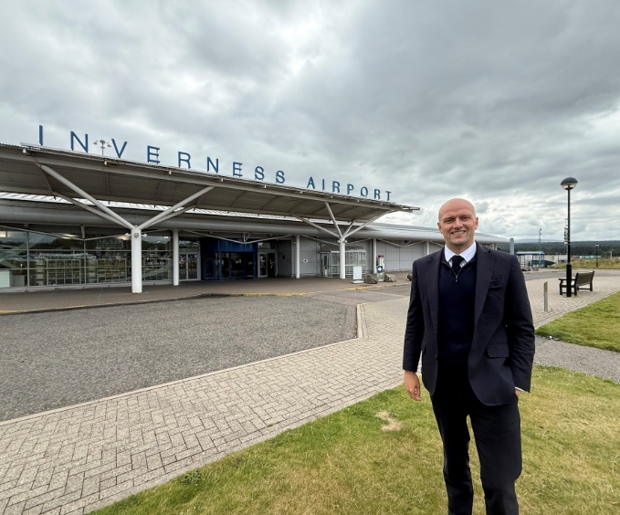 Man in formal attire standing on grass outside Inverness Airport under a cloudy sky.