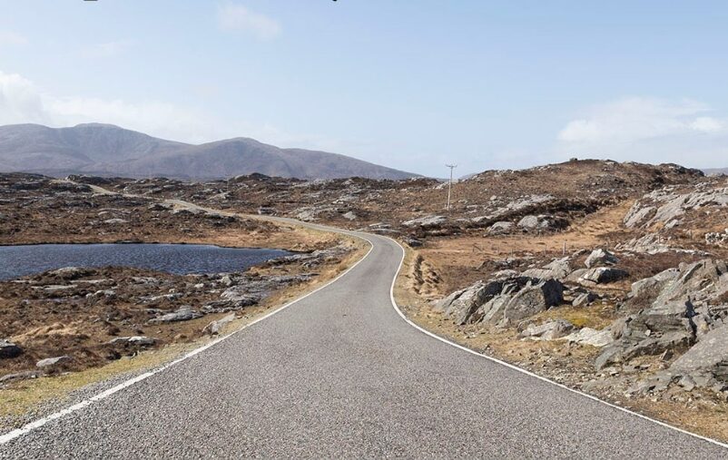 Winding road cutting through a rocky, barren landscape with distant hills under a clear blue sky.