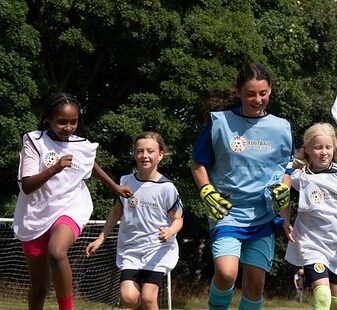 Four young girls running on a sport field during practice with a goal and footballs in the background.