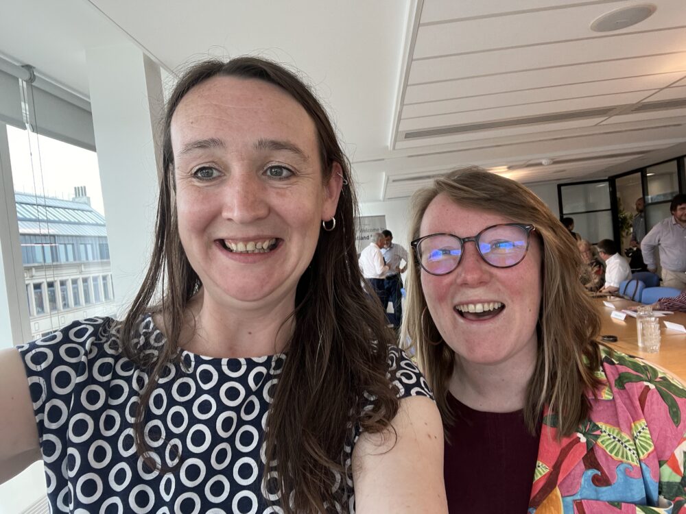 Two women smiling and posing for a selfie in a bright office space with large windows and colleagues in the background.