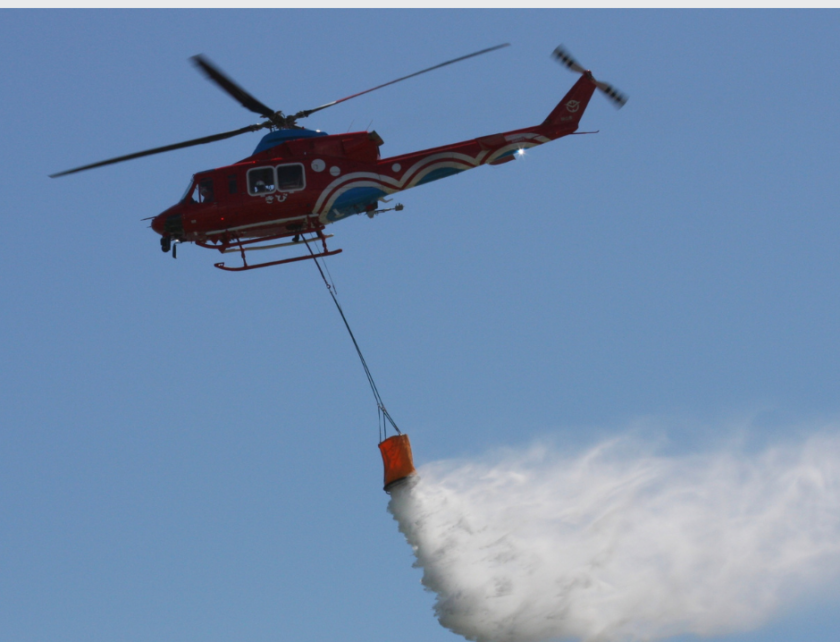 Red helicopter releasing water from a suspended bucket against a clear blue sky during firefighting operations.