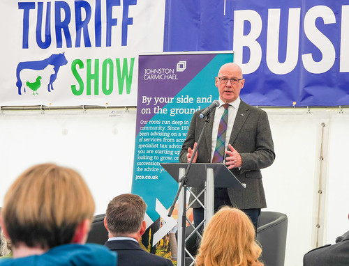 John Swinney, First Minister of Scotland in a suit and tartan tie speaks at a podium during the Turriff Show business event with an audience in front of him.