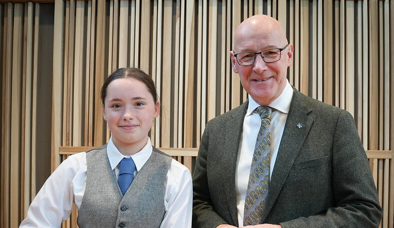 Young girl in traditional Scottish attire holding bagpipes stands next to a smiling older man in a suit and tie against a wooden panel background.