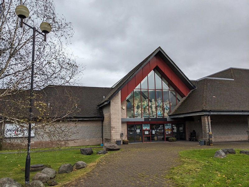 Entrance of a modern building with a large triangular glass facade and a mural above the doors under a cloudy sky.