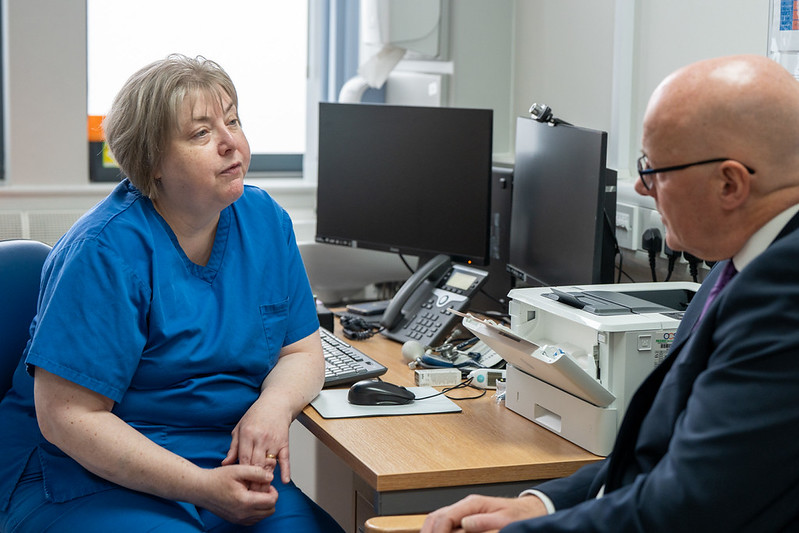 Nurse in blue scrubs attentively listening to a man in a suit during a discussion in a medical office.