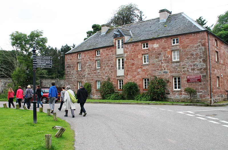 Group of people walking near a large historic red brick building surrounded by greenery on a cloudy day.