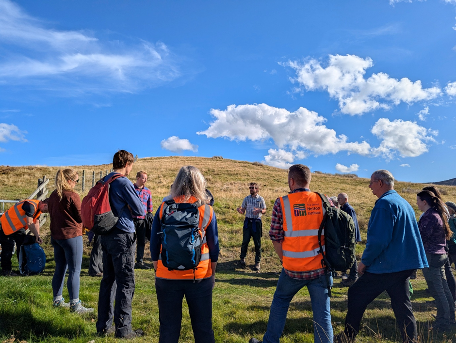 Group gathered on a grassy hill listening to a guide under a bright blue sky with scattered clouds.