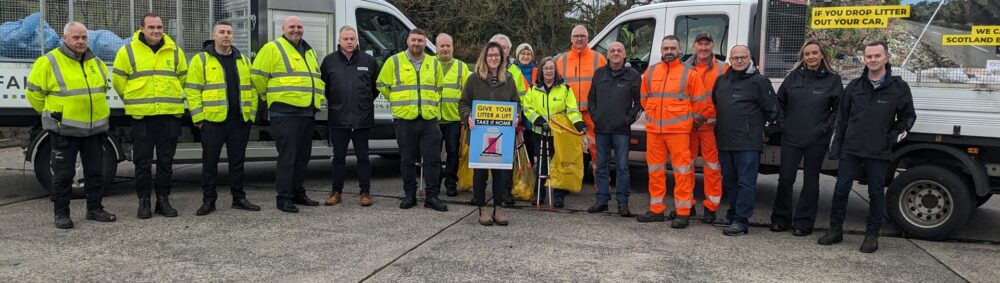 Group of workers and officials in high-visibility jackets standing in front of service vehicles during a community litter campaign event.