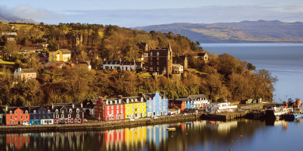 Colourful row of houses along a calm waterfront with hills and trees in the background under a clear sky.