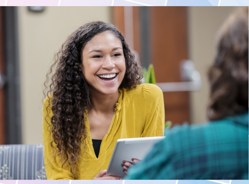 Young woman with curly hair in a yellow blouse smiling while holding a tablet during a conversation indoors.