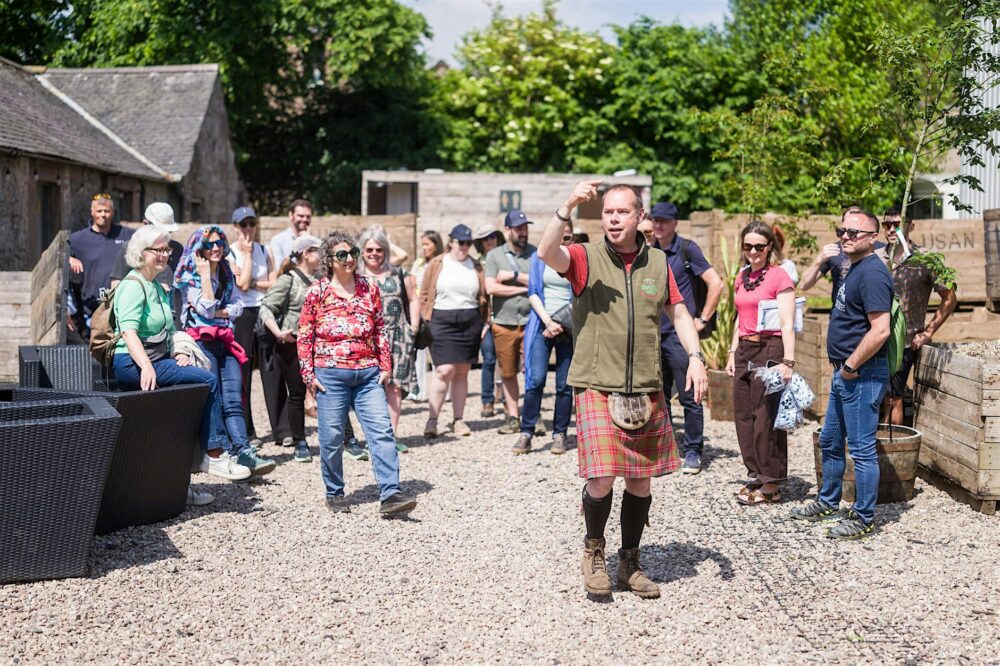 Tour guide in a kilt addressing a group of visitors outdoors on a sunny day near rustic stone buildings.