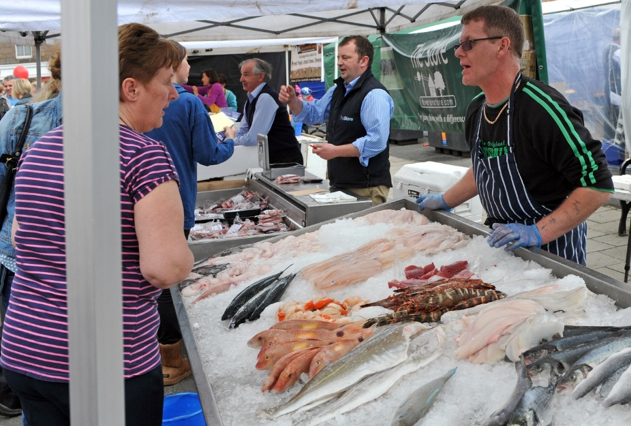 Fishmonger serving customers at an outdoor market stall with a wide variety of fresh fish and seafood displayed on ice.