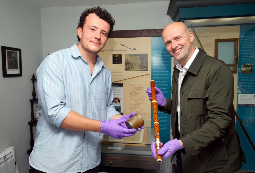 Two men wearing purple gloves examine historical artifacts in a museum exhibit room with informational displays behind them.