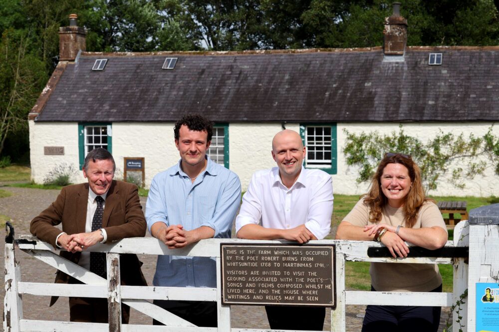 Four people smiling and leaning on a white wooden gate in front of a historic white cottage with a slate roof and green trim.