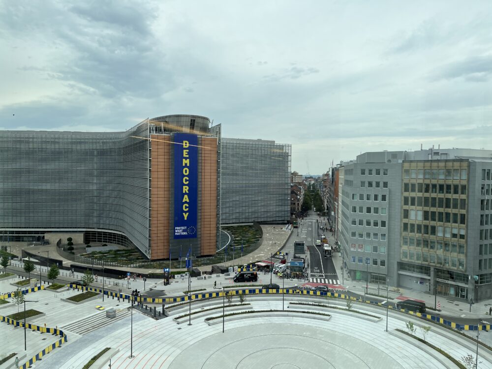 Modern curved building with a large vertical banner reading 'DEMOCRACY' in blue and yellow, overlooking a busy urban roundabout.