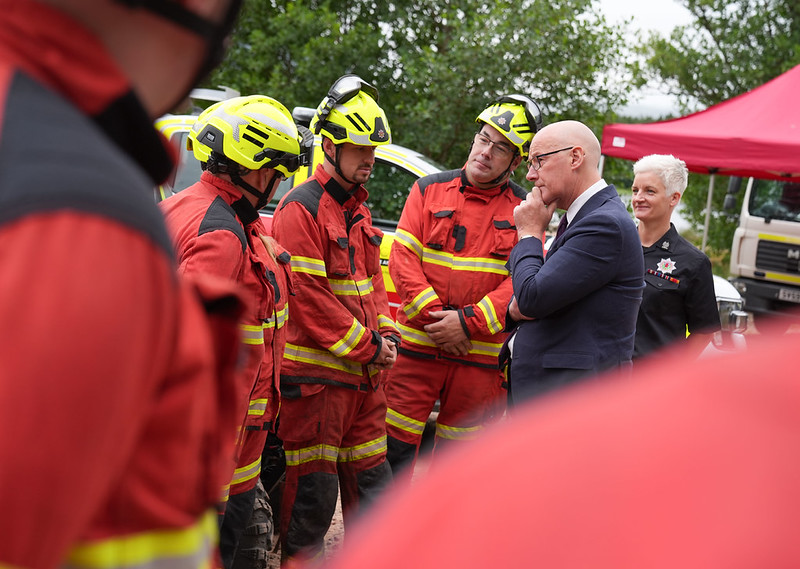 Group of firefighters in red and yellow gear speaking to a man in a suit during an outdoor briefing.