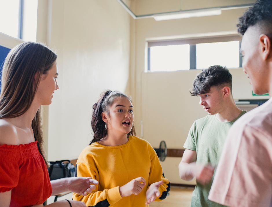 Four young adults engaged in an animated discussion inside a bright room with large windows.