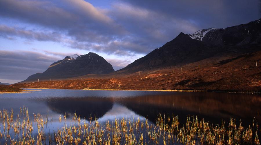 Calm loch reflecting rugged mountains under a moody, cloud-filled sky at dusk.