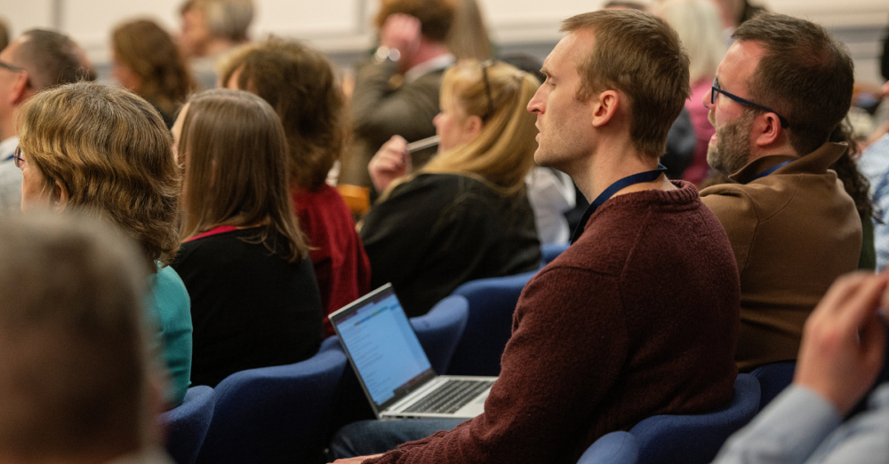 Audience members attentively listening during a conference session, with one person using a laptop to take notes.