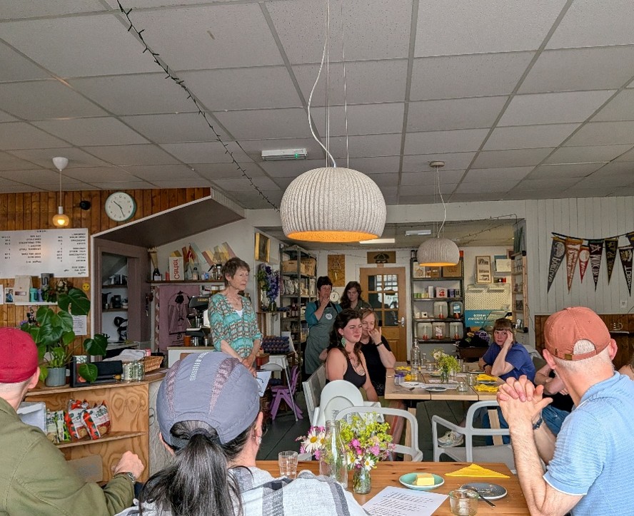 A cafe scene with people seated around tables, listening to a woman standing and speaking near the counter area.
