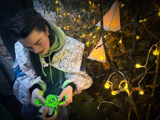 Young woman in a patterned jacket holding a glowing green object, surrounded by warm fairy lights on tree branches at night.
