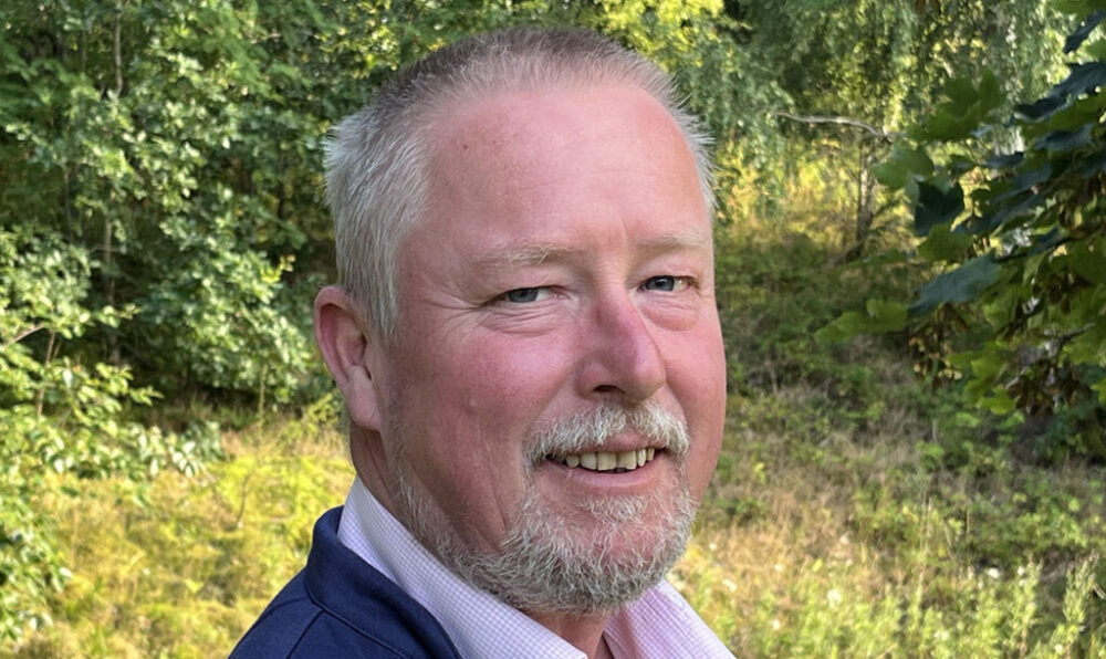 Man with short grey hair and beard smiling outdoors in a sunlit green forest setting.