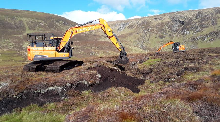 Two orange excavators digging peat soil in a rugged, grassy hillside under a partly cloudy sky.