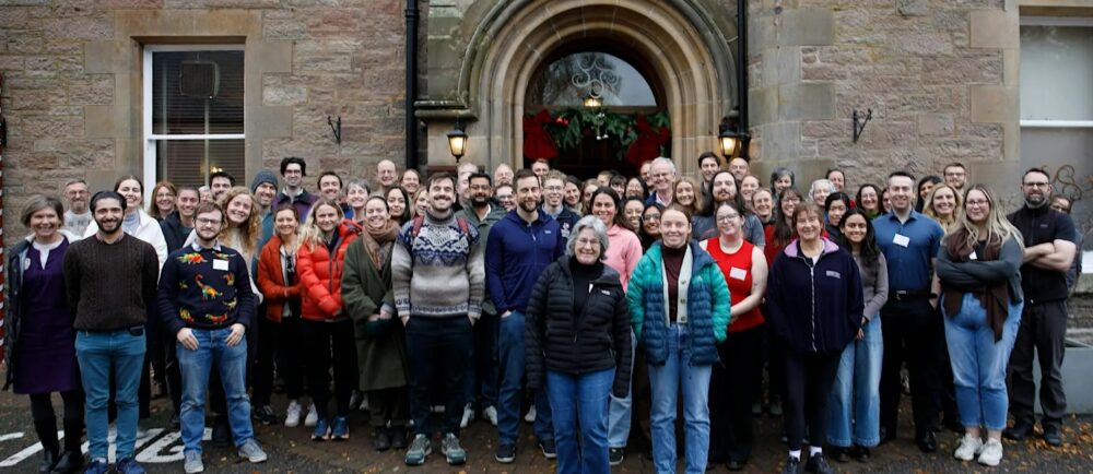 Large group of diverse adults standing outside a stone building with an arched doorway, smiling at the camera on a cloudy day.