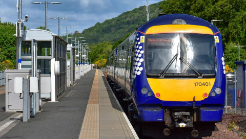 Blue and yellow passenger train stopped at a rural station platform with green hills in the background.