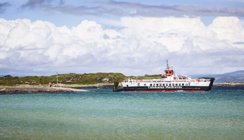 Caledonian MacBrayne ferry sailing on calm blue waters near a green, rocky island under a cloudy sky.