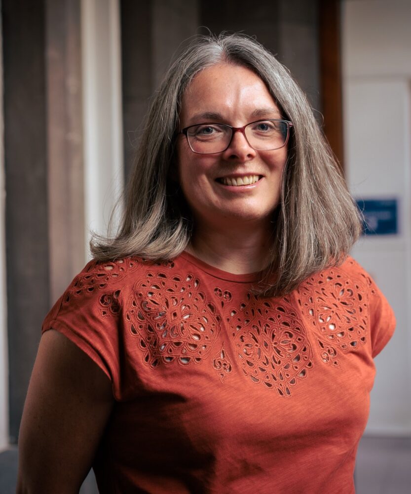 Portrait of a woman with shoulder-length grey hair and glasses in a detailed orange top.