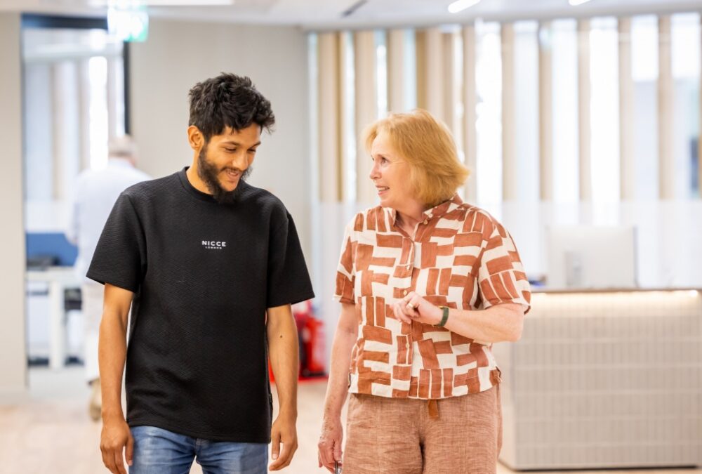 Young man and older woman walking and talking in a bright modern office space with large windows.