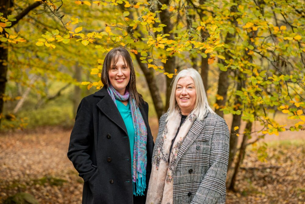 Women dressed in warm coats standing side by side under trees with bright yellow leaves.