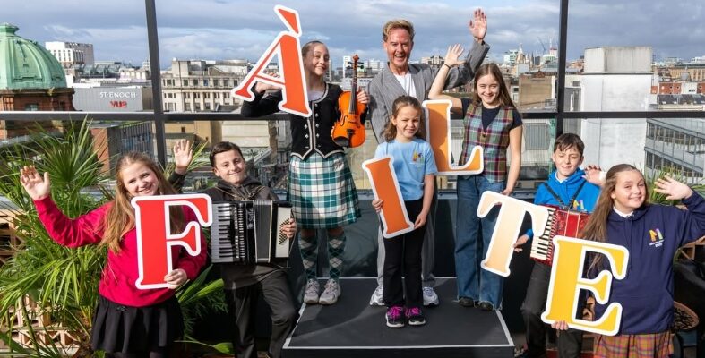 Group of children and an adult on a rooftop holding large colorful letters spelling 'FÀILTE' with city buildings in the background.