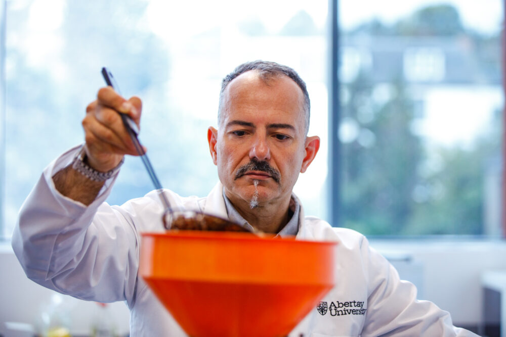 Scientist in a white lab coat carefully examining a sample using a tool over an orange funnel in a bright laboratory.